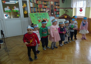 Dzieci z opaskami na głowie stoją w rzędzie i recytują wiersz. Za nimi plakat "Nie pal przy mnie proszę".